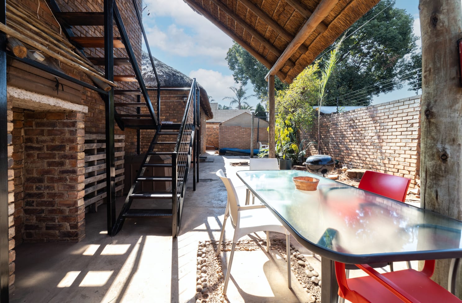 Outdoor dining area under a thatch awning with a glass table and view of the braai facilities and garden stairs.