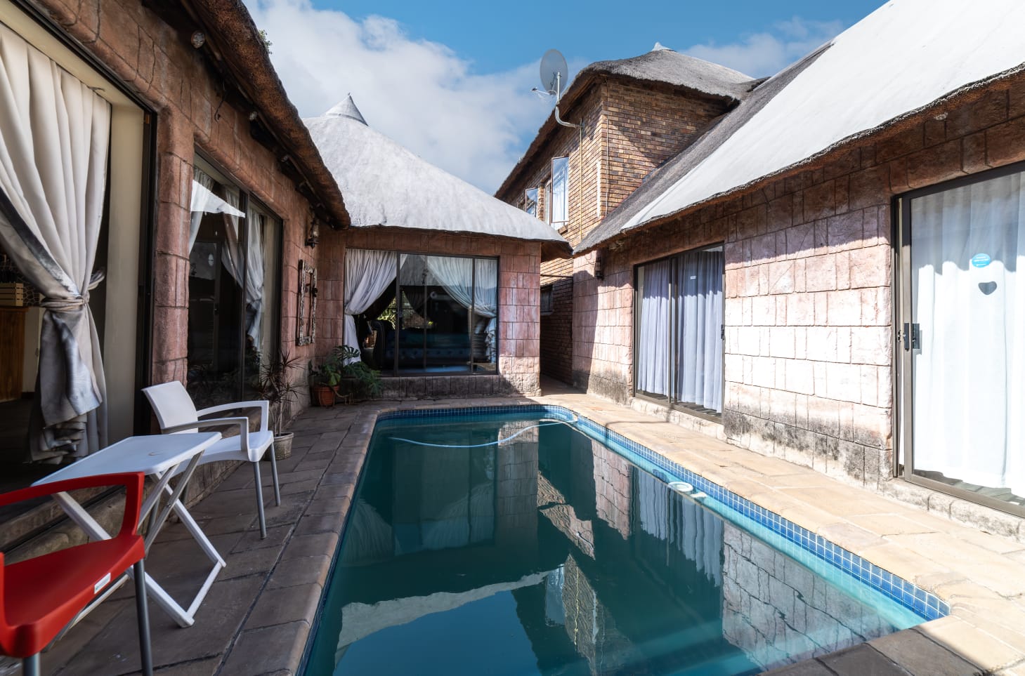 Outdoor dining area under a thatch awning with a glass table and view of the braai facilities and garden stairs.